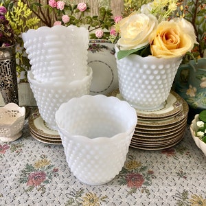 May include: A collection of white hobnail vases with scalloped rims. Some vases are stacked, while others hold yellow and white roses. The vases sit on decorative plates and a floral patterned tablecloth.