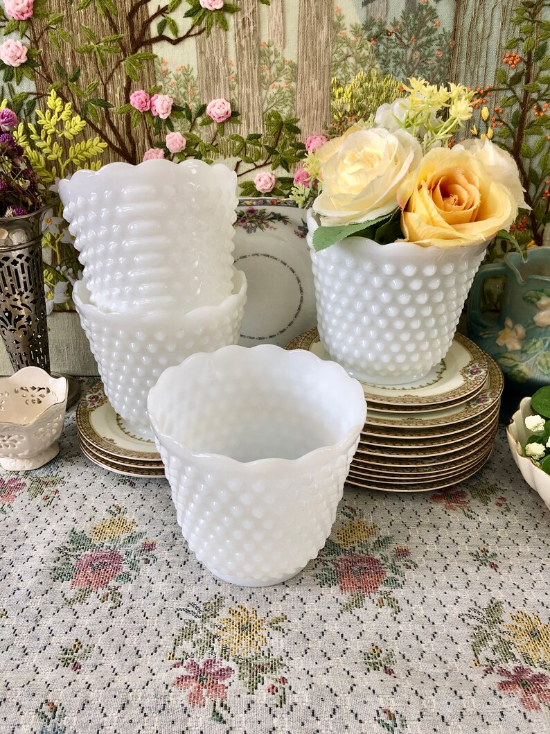 May include: A collection of white hobnail vases with scalloped rims. Some vases are stacked, while others hold yellow and white roses. The vases sit on decorative plates and a floral patterned tablecloth.