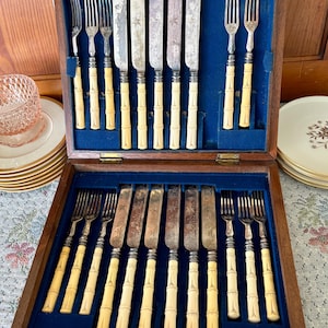 Pode incluir: Uma caixa de talheres de madeira aberta com interior de veludo azul, contendo um conjunto de facas e garfos vintage. As al&ccedil;as s&atilde;o de cor creme, projetadas para se assemelhar ao bambu. As l&acirc;minas das facas mostram sinais de idade.