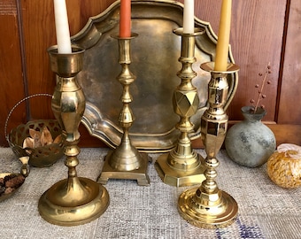 May include: Four gold metal candle holders with different designs, each holding a candle. The candles are white, orange, and yellow. The candle holders are on a gray and white patterned rug.