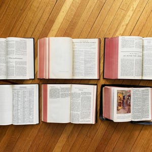 May include: Six open books with black covers and red edges. The books are laid out on a wooden floor. The titles of the books are visible: "I Thessalonians", "Scripture Proper Names", "St. Peter the Apostle", "Titus", and "The Acts of the Apostles".