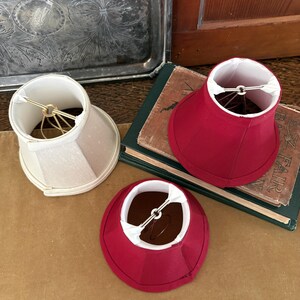 Op de afbeelding: Drie kleine lampenkappen. E&eacute;n is gebroken wit, de andere twee zijn dieprood. De kappen liggen op een bruin oppervlak naast een stapel boeken met een groene kaft. De kappen hebben een goudkleurig draadframe.