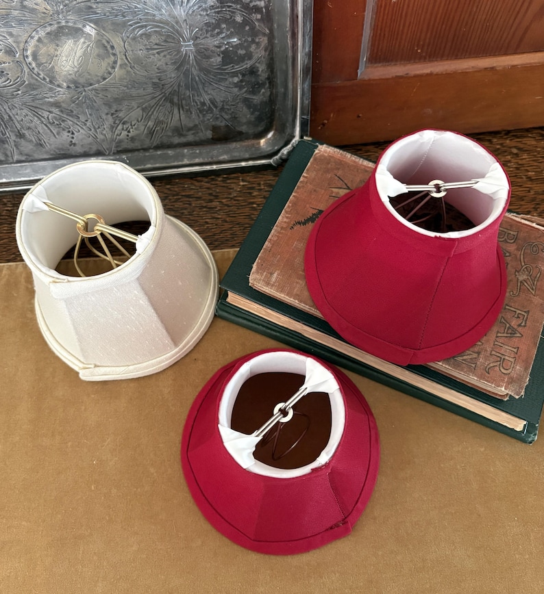 Op de afbeelding: Drie kleine lampenkappen. E&eacute;n is gebroken wit, de andere twee zijn dieprood. De kappen liggen op een bruin oppervlak naast een stapel boeken met een groene kaft. De kappen hebben een goudkleurig draadframe.