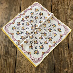 K&ouml;nnte beinhalten: Ein Vintage-Taschentuch mit floralem Muster. Der wei&szlig;e Stoff ist mit gelben Rosen, lila Akzenten und gr&uuml;nen Bl&auml;ttern verziert. Es hat einen gelben Rand und dekoratives lilafarbenes Netz an den R&auml;ndern.