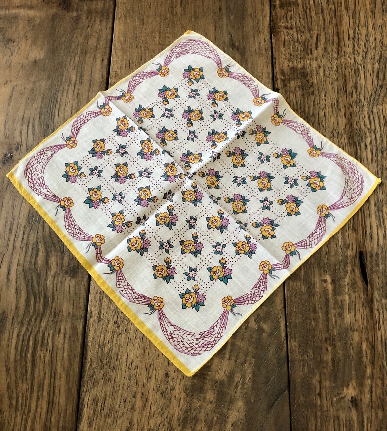 K&ouml;nnte beinhalten: Ein Vintage-Taschentuch mit floralem Muster. Der wei&szlig;e Stoff ist mit gelben Rosen, lila Akzenten und gr&uuml;nen Bl&auml;ttern verziert. Es hat einen gelben Rand und dekoratives lilafarbenes Netz an den R&auml;ndern.