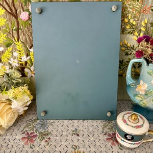May include: A rectangular, teal-colored picture frame with four small, gray pegs in each corner. The frame is displayed on a floral patterned surface, with a decorative teacup and a blue pitcher in the background.