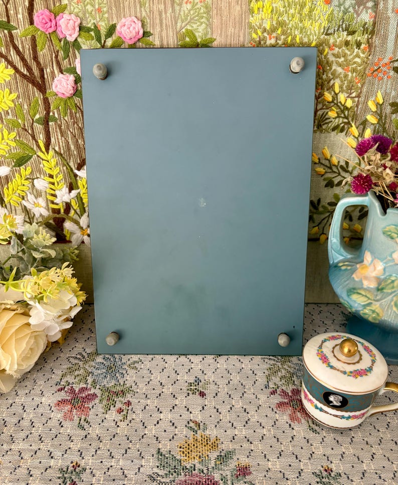 May include: A rectangular, teal-colored picture frame with four small, gray pegs in each corner. The frame is displayed on a floral patterned surface, with a decorative teacup and a blue pitcher in the background.