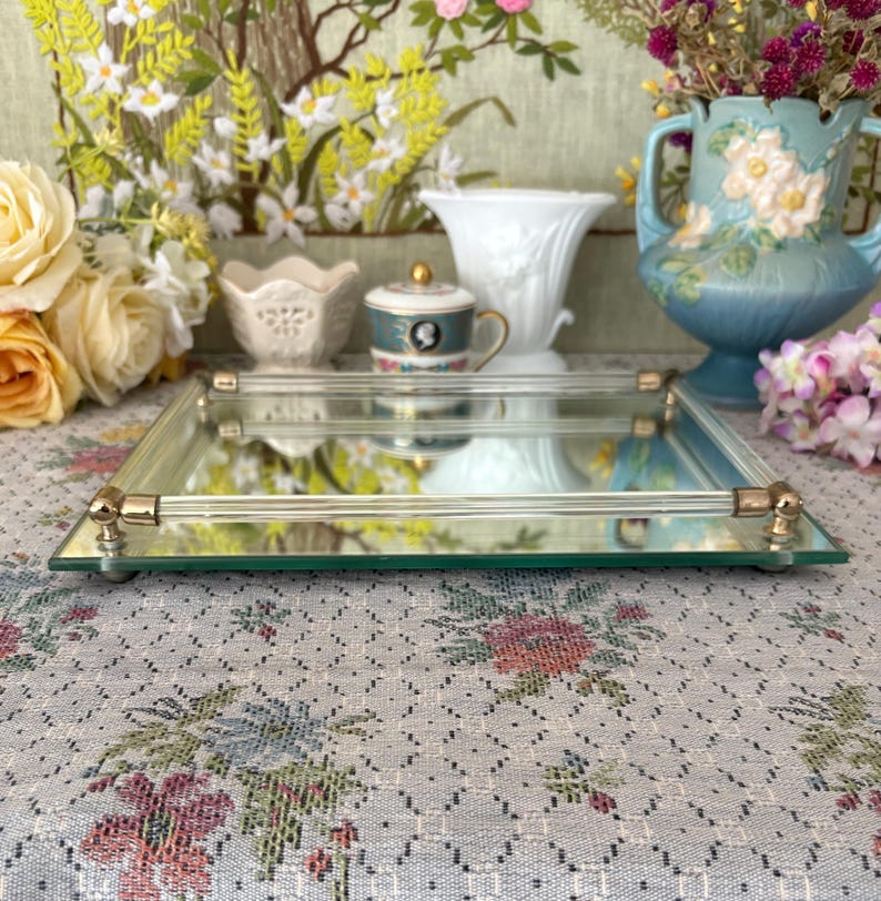 May include: A rectangular mirrored tray with gold-colored accents. The tray has a clear glass surface and is surrounded by a gold-colored metal frame. The tray is decorated with small gold-colored feet and is surrounded by decorative items.