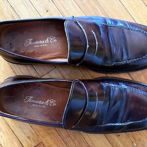 May include: A pair of dark brown leather penny loafers with a polished finish and visible stitching along the toe. The interior insoles are stamped with the brand name Framosca & Co and the text Made in Italy in a gold-colored script font.
