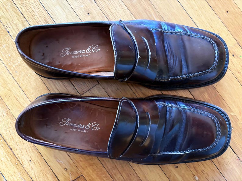 May include: A pair of dark brown leather penny loafers with a polished finish and visible stitching along the toe. The interior insoles are stamped with the brand name Framosca & Co and the text Made in Italy in a gold-colored script font.