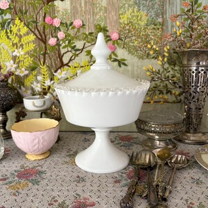 May include: A white, lidded pedestal candy dish with a decorative rim. The dish is made of opaque glass and sits on a round base. The background includes a floral tapestry, a pink bowl, and antique silverware.