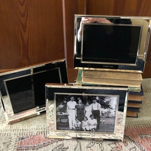 May include: Three silver picture frames with black mats. The frame on the left is empty. The middle frame has a black and white photo of a family. The frame on the right is empty. The frames are on a stack of books.