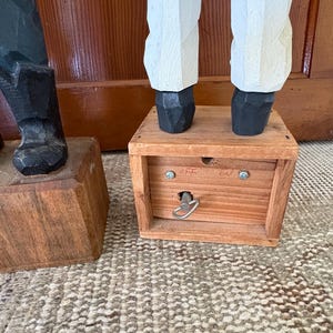 May include: Wooden carved figure of a sailor with white pants, black shoes, and a blue jacket. The figure stands on a wooden box with a key and the words "off" and "on" are visible.