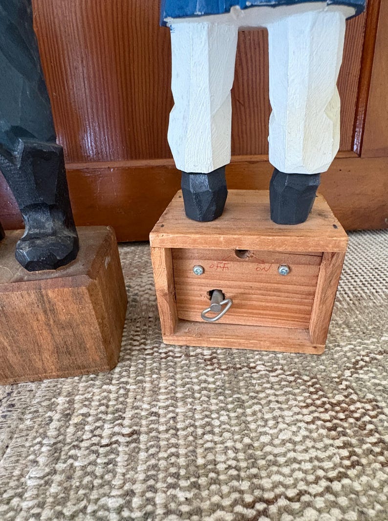 May include: Wooden carved figure of a sailor with white pants, black shoes, and a blue jacket. The figure stands on a wooden box with a key and the words "off" and "on" are visible.