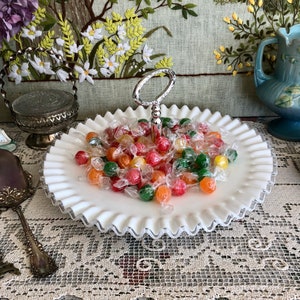 Puede incluir: Un plato de caramelo de vidrio de leche blanco con un asa plateada y un borde rizado. El plato est&aacute; lleno de caramelos coloridos.