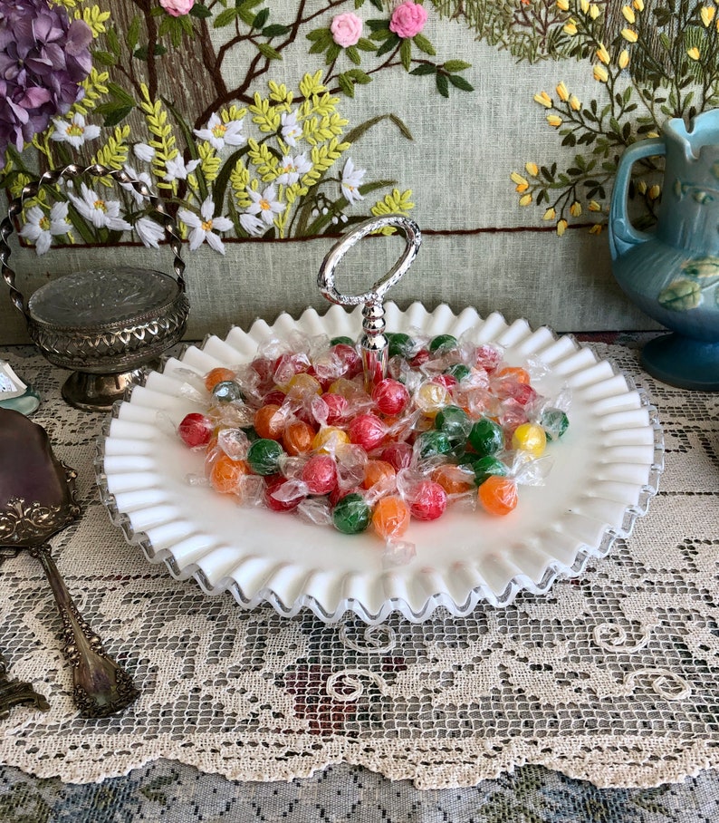 Puede incluir: Un plato de caramelo de vidrio de leche blanco con un asa plateada y un borde rizado. El plato est&aacute; lleno de caramelos coloridos.