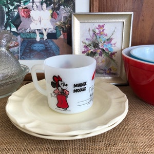 May include: White ceramic mug with a red and white image of Minnie Mouse. The mug has the text "Minnie Mouse" on it. The mug is sitting on a white saucer.