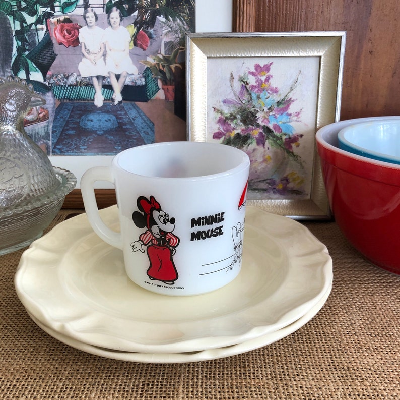May include: White ceramic mug with a red and white image of Minnie Mouse. The mug has the text "Minnie Mouse" on it. The mug is sitting on a white saucer.