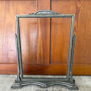May include: An antique, rectangular, metal picture frame with a decorative top and base. The frame is supported by two angled side supports. The frame has a distressed, aged appearance with a muted, metallic finish.