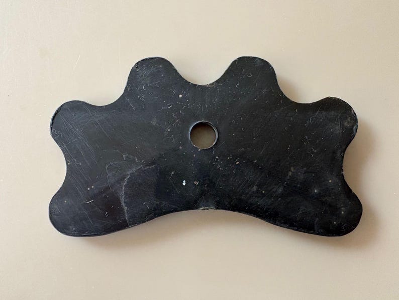 K&ouml;nnte beinhalten: Ein schwarzer Billardqueue-Br&uuml;ckenhalter mit gewelltem Rand und einem zentralen Loch. Der Br&uuml;ckenhalter besteht aus einem soliden, dunklen Material und hat eine glatte, leicht abgenutzte Oberfl&auml;che. Das Design ist symmetrisch, mit drei gebogenen Abschnitten auf jeder Seite.
