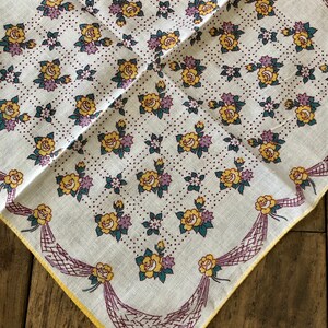 K&ouml;nnte beinhalten: Quadratisches wei&szlig;es Taschentuch mit floralem Muster. Das Design zeigt gelbe Rosen, lila Blumen und gr&uuml;ne Bl&auml;tter, die in einem Gittermuster angeordnet sind. Die R&auml;nder sind mit einem gelben Rand und dekorativen lila B&auml;ndern versehen.