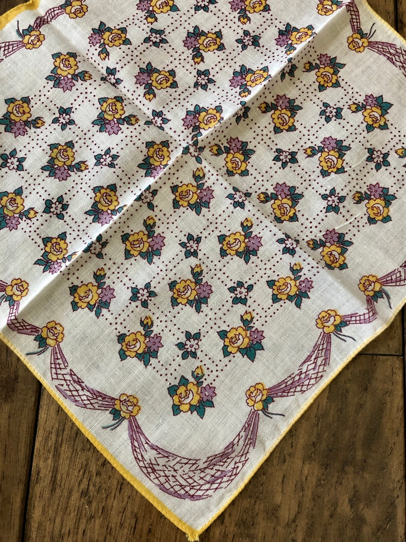 K&ouml;nnte beinhalten: Quadratisches wei&szlig;es Taschentuch mit floralem Muster. Das Design zeigt gelbe Rosen, lila Blumen und gr&uuml;ne Bl&auml;tter, die in einem Gittermuster angeordnet sind. Die R&auml;nder sind mit einem gelben Rand und dekorativen lila B&auml;ndern versehen.