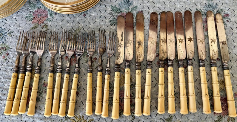 Pode incluir: Uma cole&ccedil;&atilde;o de talheres antigos, incluindo garfos e facas. Os garfos e facas t&ecirc;m cabos de cor clara com um design de bambu. As l&acirc;minas das facas t&ecirc;m um design decorativo. Os talheres est&atilde;o dispostos sobre uma toalha de mesa com padr&atilde;o floral.