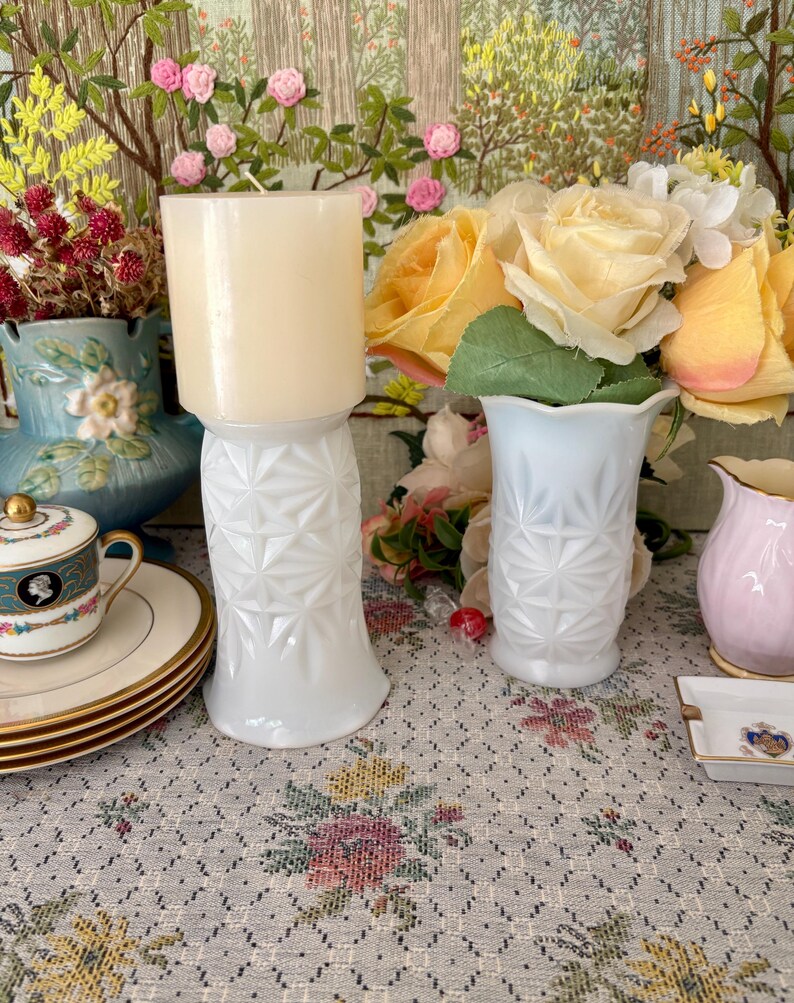 Pu&ograve; includere: Due vasi in vetro opalino bianco con un motivo geometrico a stella. Un vaso contiene una candela cilindrica color crema, mentre l'altro ospita un bouquet di rose artificiali gialle e bianche.