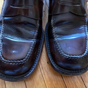 May include: A pair of dark burgundy leather penny loafers featuring a polished finish, a classic moc-toe construction with visible stitching along the vamp, and a traditional strap across the top of each shoe, showcasing a smooth, glossy texture.
