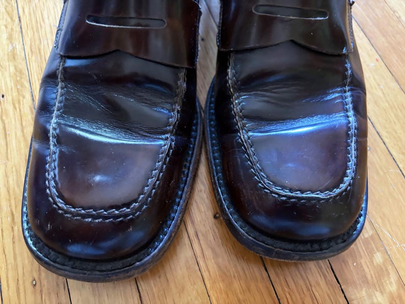 May include: A pair of dark burgundy leather penny loafers featuring a polished finish, a classic moc-toe construction with visible stitching along the vamp, and a traditional strap across the top of each shoe, showcasing a smooth, glossy texture.