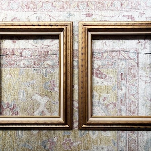 May include: A pair of rectangular picture frames featuring a metallic gold finish with a stepped, tiered profile. Each frame includes a thin, light-coloured inner border and a thin metal hanging wire attached to the back for wall mounting purposes.