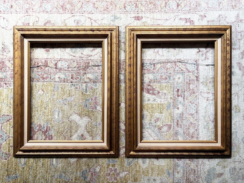 May include: A pair of rectangular picture frames featuring a metallic gold finish with a stepped, tiered profile. Each frame includes a thin, light-coloured inner border and a thin metal hanging wire attached to the back for wall mounting purposes.