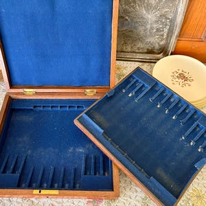 Pode incluir: Caixa de armazenamento de talheres de madeira aberta com interior de feltro azul. A caixa &eacute; retangular com compartimentos para utens&iacute;lios. Uma pilha de pratos com um desenho floral &eacute; vis&iacute;vel no fundo.