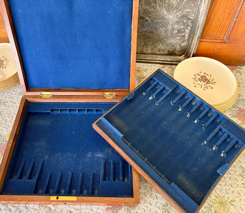 Pode incluir: Caixa de armazenamento de talheres de madeira aberta com interior de feltro azul. A caixa &eacute; retangular com compartimentos para utens&iacute;lios. Uma pilha de pratos com um desenho floral &eacute; vis&iacute;vel no fundo.