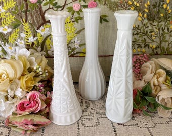 May include: Three white vintage milk glass vases of varying heights and designs. The vases are arranged on a lace tablecloth, with floral arrangements in the foreground and a floral tapestry in the background. The vases have textured surfaces.