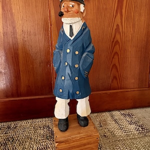 May include: A hand-carved wooden nautical captain figurine. The captain wears a navy blue coat with gold buttons, white pants, and a white hat. He has a pipe in his mouth and stands on a small wooden box. The figurine is a decorative item.