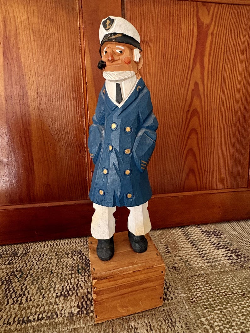May include: A hand-carved wooden nautical captain figurine. The captain wears a navy blue coat with gold buttons, white pants, and a white hat. He has a pipe in his mouth and stands on a small wooden box. The figurine is a decorative item.