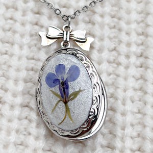 Op de afbeelding: Zilveren ketting met een delicaat strikbedeltje. Het medaillon heeft een geperste bloem, een lichtblauwe bloem met groene stelen, ingekapseld in helder hars.