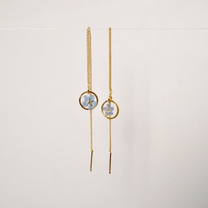 Puede incluir: Un par de pendientes de cadena dorada con una delicada flor azul encerrada en un colgante circular de oro.