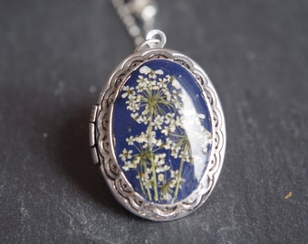 May include: Oval silver locket pendant with a dark blue background, showcasing delicate white dried flowers. The locket is on a silver chain, and the edges have a decorative scalloped design.