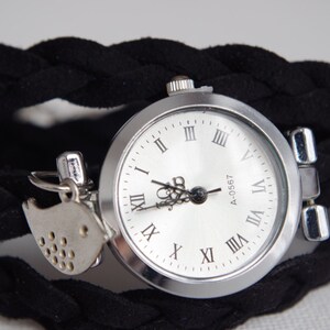 Peut inclure: Une montre argentée avec un cadran blanc et des chiffres romains noirs. La montre est attachée à un bracelet en tissu noir tressé. Un petit charme d'oiseau argenté pend à la montre.