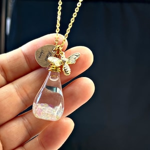 Puede incluir: Collar de oro con un pequeño colgante de botella de vidrio lleno de purpurina iridiscente. La botella está unida a un dije de abeja de oro y un dije ovalado de oro con la inscripción "Mama, eres mágica".