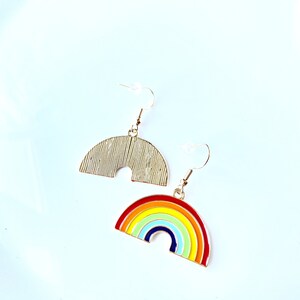 May include: A pair of gold-tone earrings featuring a rainbow design. One earring is a solid gold arch, while the other is a rainbow arch with red, orange, yellow, green, blue, and purple stripes.