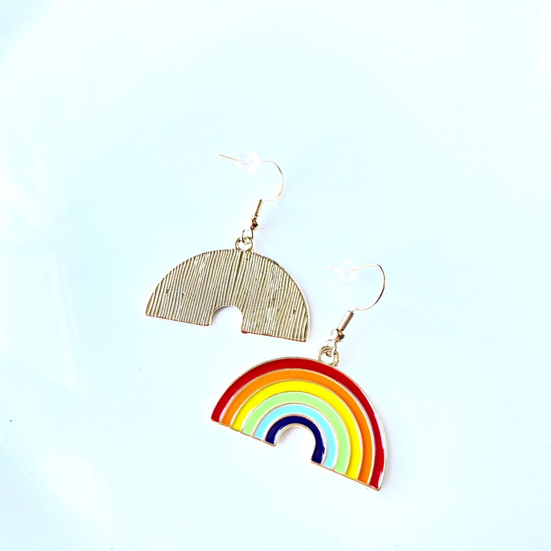 May include: A pair of gold-tone earrings featuring a rainbow design. One earring is a solid gold arch, while the other is a rainbow arch with red, orange, yellow, green, blue, and purple stripes.