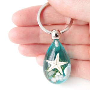 Op de afbeelding: Een turquoise glazen traanvormige sleutelhanger met een witte zeester, witte schelpen en groen zeewier erin. De sleutelhanger is bevestigd aan een zilveren metalen ring.