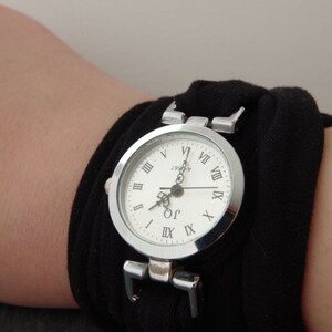 K&ouml;nnte beinhalten: Eine silberne Armbanduhr mit r&ouml;mischen Ziffern auf einem wei&szlig;en Zifferblatt. Die Uhr ist an einem schwarzen Stoffband befestigt.