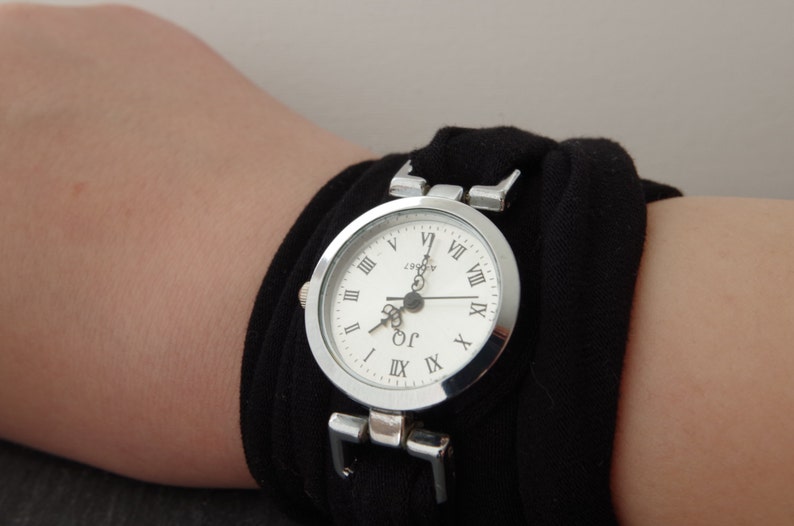 K&ouml;nnte beinhalten: Eine silberne Armbanduhr mit r&ouml;mischen Ziffern auf einem wei&szlig;en Zifferblatt. Die Uhr ist an einem schwarzen Stoffband befestigt.