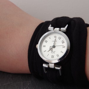 K&ouml;nnte beinhalten: Eine silberne Armbanduhr mit r&ouml;mischen Ziffern auf einem wei&szlig;en Zifferblatt. Die Uhr ist an einem schwarzen Stoffband befestigt.