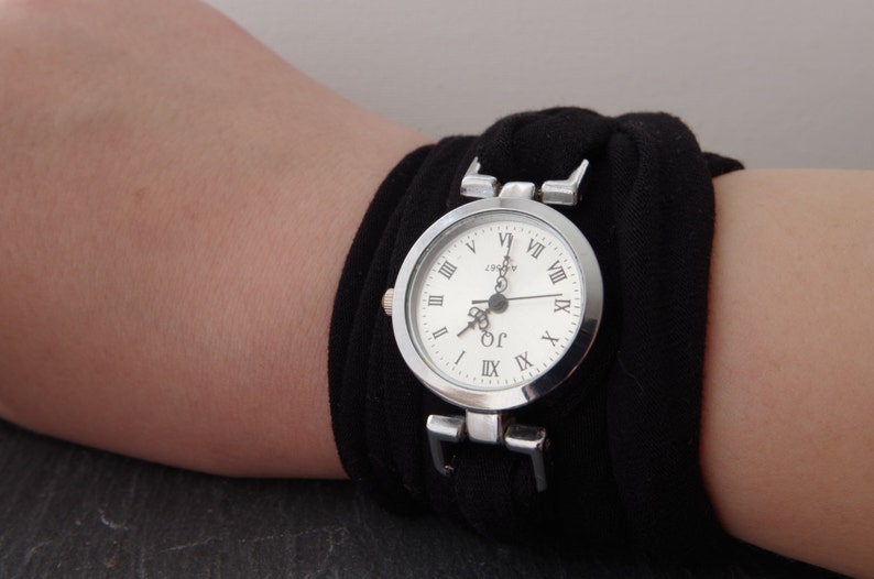 K&ouml;nnte beinhalten: Eine silberne Armbanduhr mit r&ouml;mischen Ziffern auf einem wei&szlig;en Zifferblatt. Die Uhr ist an einem schwarzen Stoffband befestigt.