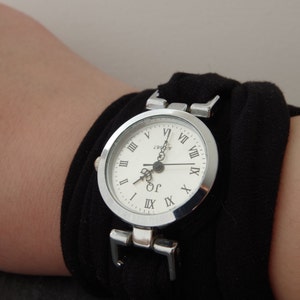 K&ouml;nnte beinhalten: Eine silberne Armbanduhr mit r&ouml;mischen Ziffern auf einem wei&szlig;en Zifferblatt. Die Uhr wird an einem schwarzen Stoffarmband getragen.